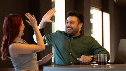 Middle-aged businessman and young businesswoman celebrating enthusiastically and high-fiving each other during a successful meeting with laptops at a café outside the office