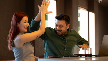 Middle-aged businessman and young businesswoman celebrating enthusiastically and high-fiving each other during a successful meeting with laptops at a café outside the office