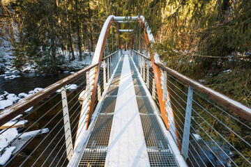 Obraz premium Snow-covered suspension bridge with metal railings crossing the Ahja River in the picturesque winter forest of Taevaskoja, Estonia, on a clear sunny day.