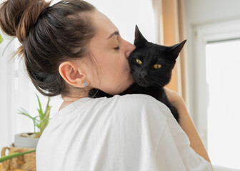 Happy young beautiful woman kisses and holds a black cat. Playing with a pet at home. Love, comfort, relaxation, animal protection concept.
