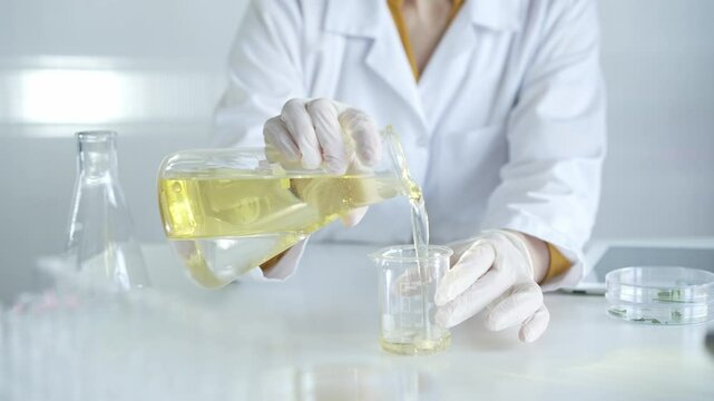 Unrecognizable woman scientist, wearing a lab coat and white protective gloves, is pouring a yellow oily liquid from one beaker to another in laboratory, close up. Medicine and microbiology science