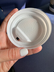Close-up of a white disposable coffee cup lid held in a hand with manicured nails. The lid on the inside is dotted with small droplets of condensation from the hot drink.