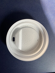Close-up of a white disposable coffee cup lid on a blue plastic table. The inside of the lid is dotted with small droplets of condensation from the hot drink. 