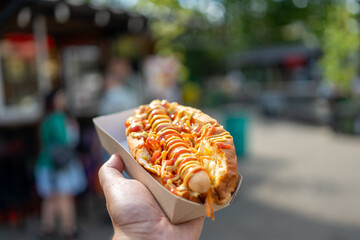 A close-up of a hand holding a delicious hot dog topped with ketchup and mustard, set against a vibrant outdoor food market background, showcasing a lively atmosphere.