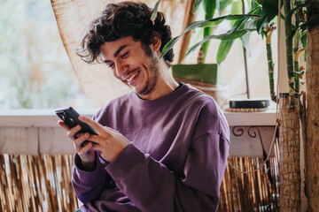 A cheerful young man relaxes indoors, smiling while using his smart phone. The setting is cozy,...
