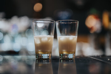 Two cocktail glasses filled with an iced drink rest on a bar with blurred lights in the background, encapsulating the atmosphere of a bar or social gathering.