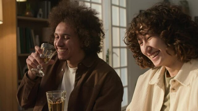 Man and woman with curly hair clinking glasses of light-colored drink in sunlit room at dinner party