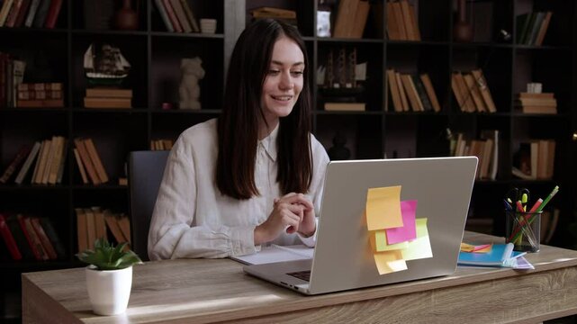 Young smiling brunette woman participates in an online interview and enthusiastically answers questions. Woman sits at an office table and talks in front of a computer explaining with her hands