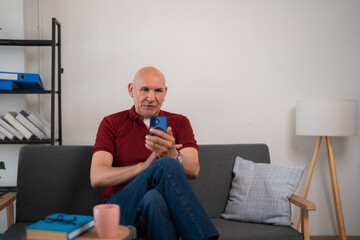 Fototapeta premium Bald senior man using mobile phone sitting on sofa at home, browsing internet or reading messages, modern communication and lifestyle concept