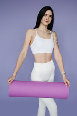 brunette woman yoga trainer smiling and holding a yoga mat, purple background 