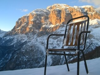 An empty metal chair surrounded by snow sits in the foreground, framed by majestic snow-capped mountains illuminated by the warm glow of sunset. Nature escape.