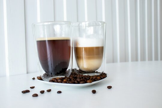 Double-walled glasses filled with espresso and cappuccino on a white plate with coffee beans