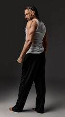 Fototapeta premium Confident bearded man with long hair wearing white tank top and black pants, posing against dark studio background. Side view. Concept of male beauty, self-care, masculinity.
