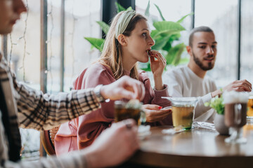 A diverse group of friends gather at a cafe, enjoying a casual meal, drinks, and each other's company in a cozy atmosphere with a backdrop of natural lighting and greenery.