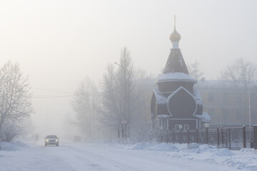Winter landscape with snow-covered street and Orthodox church. Car is driving along the road. Very cold weather. Winter frost. Frosty fog. City of Kyzyl, Republic of Tyva (Tuva), Siberia, Russia.