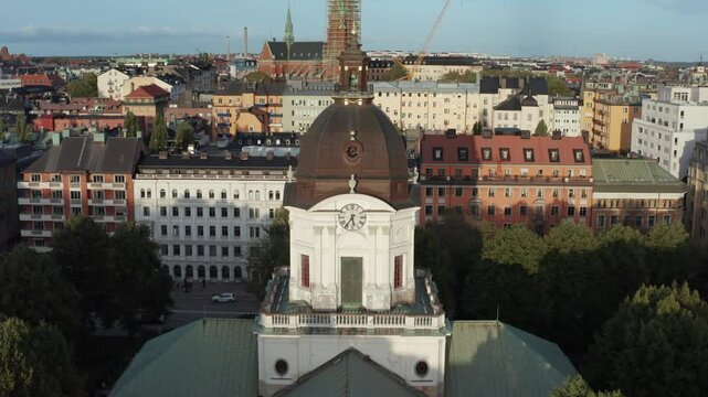 Drone shot of Adolf Fredriks church in Stockholm, Sweden during sunset in late summer. Drone rising upwards tilting down with church in foreground revealing buildings and Sveav&auml;gen in background.