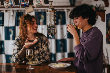 Two people are sitting indoors, engaging in conversation while sharing dinner and beverages. The setting appears cozy and inviting, showcasing friendship and warm interactions in a relaxed atmosphere.