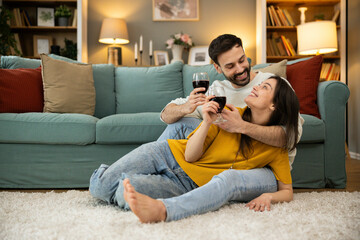 Obraz premium A joyful couple share a special moment while sitting on the floor, enjoying a glass of wine in a warm, cozy living room setting.