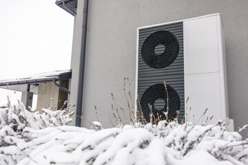 Close-up of a dual-fan outdoor heat pump mounted on a gray wall, surrounded by snow-covered bushes...