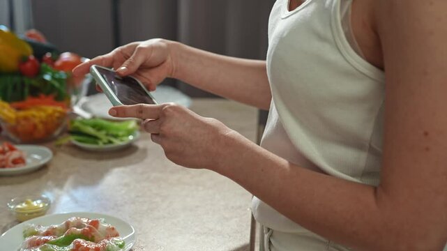 Young woman at the table in the kitchen preparing spring rolls for healthy lunch, takes pictures on smartphone of plate with fresh spring rolls. Slow motion.