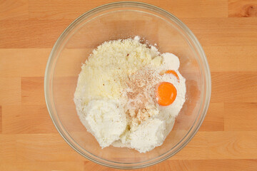 A top-down view of a glass bowl filled with ingredients for cheese and flour gnocchi preparation, including flour, cheese, egg, and spices, on a wooden surface