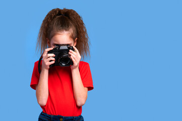 Girl in red T-shirt holds camera and takes shot against blue background. Young photographer...