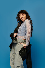 Stylish young woman with curly hair poses confidently in trendy urban outfit against blue backdrop