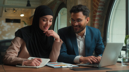 Male female teamwork Arabian man muslim woman in hijab girl guy businessman businesswoman two happy business colleagues coworkers managers analyzing marketing graphs documents in office typing laptop