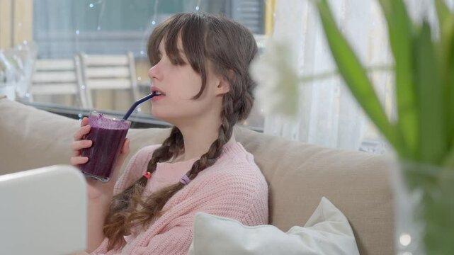 A teenage girl in a pink knitted sweater is drinking a freshly blended berry smoothie from a glass. Healthy nutrition, homemade drinks, and a balanced diet for kids and teenagers