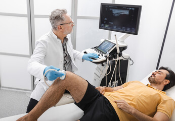 Medical expert in white coat performs ultrasound on male patient's knee, evaluating injury during...