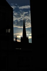 Obraz premium The tower is a tower with a high spire at the end of which a cross is seen among the buildings against the blue sky with gray clouds and bright sunlight.