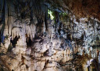 A cave with stalagmites and stalactites, illuminated with colorful lighting that highlights the natural rock formations. The textured walls and mineral structures create an ethereal atmosphere. 