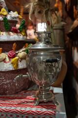 A vintage metal samovar on a traditional embroidered cloth, set in a rustic interior with decorative festive bread in the background.