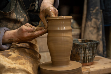A close-up of a potter’s hands covered in wet clay as they shape a tall ceramic vase on a...