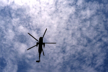 Sihouette of an unmarked helicopter flying overhead against a blue sky with white clouds
