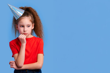 Serious girl in red T-shirt and shiny party hat crosses arms and rests chin on hand, deep in...