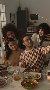 Vertical shot of four smiling friends taking selfie, leaning in for playful group photo during cozy, candlelit meal