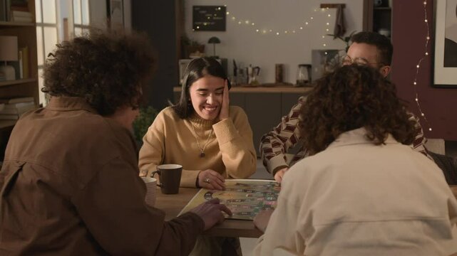 Four friends having fun when playing board game together, while sitting at table in cozy apartment