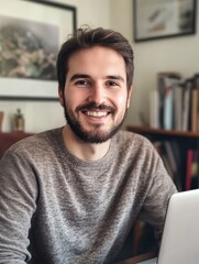 Fototapeta premium Smiling young adult bearded casual man looking at camera sitting at home office desk. Millennial guy student, freelancer, remote worker with laptop portrait. Distance job, online elearning concept.