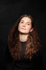 Fototapeta premium Portrait of a young woman with open eyes, curling hair in a black jacket against a black background, portrait with sunlight beam. Selective focus