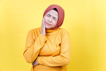 Fototapeta premium An asian woman in a hijab and yellow sweater is experiencing tooth pain, pressing her cheek with a pained expression.