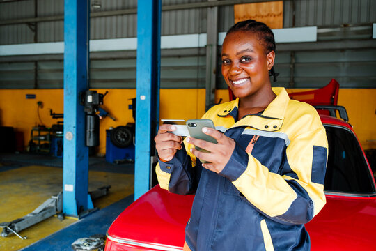 Smiling African female mechanic in a workshop using a smartphone and credit card for online payment. Concept of digital transactions, fintech, and women in the automotive industry.