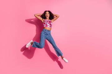 Young cheerful woman jumping joyfully against a pink background wearing stylish casual clothing...