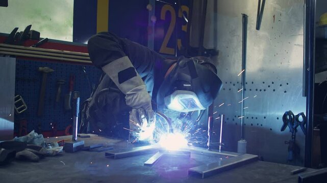 Seasoned skilled blue-collar worker working in a workshop creating sparks while welding metal materials, medium shot. Industrial concepts.