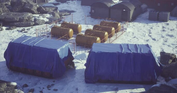 A remote research station is set in a cold, snowy landscape. Several storage containers covered in blue tarps are visible alongside yellow tanks. Rocky terrain surrounds the area.