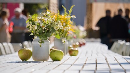 A white wooden table in the open air is decorated with bouquets of wild flowers and ripe apples. Autumn holiday, festival, outdoor food