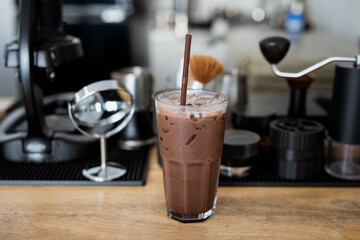 refreshing iced chocolate drink in glass with straw sits on wooden counter, surrounded by coffee making equipment in cozy cafe setting