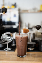 tall glass of iced chocolate drink with straw sits on wooden counter in cozy cafe setting, surrounded by coffee making equipment