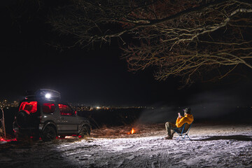 A man sits on a chair with a view of the lights of the night city. A bonfire burns with smoke. An off-road vehicle is parked on top. The concept of recreation and trips to nature. Use smartphone