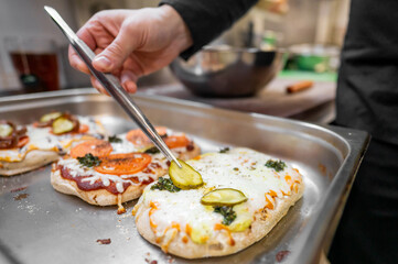 A close-up of a hand using tongs to add pickles on a freshly made pizza. The pizza is topped with melted cheese, tomatoes, and a sprinkle of herbs.